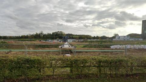 A piece of land fenced off by developers on Dame Mary Archer Way. In the foreground is a hedge and fence and beyond that metal fencing, scrubby land, rows of aggregate bags on the right. Behind them are portable cabins and on the horizon trees and sky. 