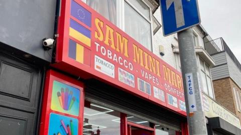 Sam Mini Market in Northampton. The building is a convenience store selling tobacco, vapes and confectionary. The branding is red with a sign with yellow writing saying SAM MINI MARKET.