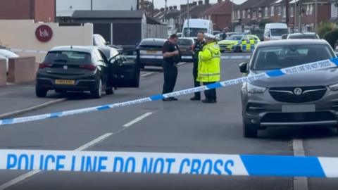 A residential street cordoned off with blue and white police tape. Three police officers, one in a hi-vis jacket, stand near a black car with its driver's door open. A police car further away is parked, blocking the road.