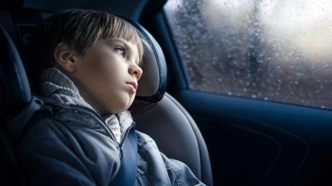 A child sits secured in a car seat in the back of a vehicle, wearing a grey coat and seatbelt, looking out through a rain-speckled side window. The interior of the car is dimly lit, with soft daylight filtering through the window and blurred scenery visible outside.