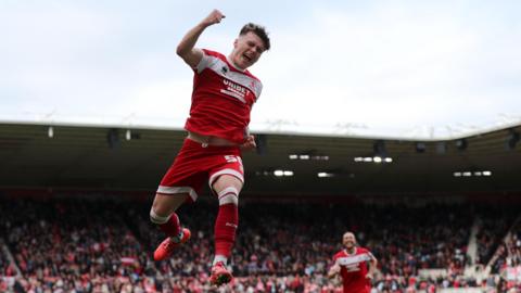 A footballer in the red shirt and shorts of Middlesbrough leaps into the air punching his fist. A teammate is running towards him smiling. In the background is a full stand of cheering fans.