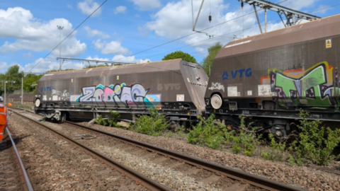 Two large freight wagons with their wheels off the tracks and overhead power lines above