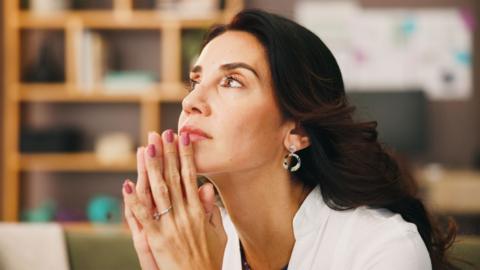 Woman in home on sofa with hands together, resting on chin and thinking