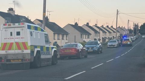 A police land rover parked at the side of the road beside a row of houses. A number of other cars are parked further along the road, one of them a police car.