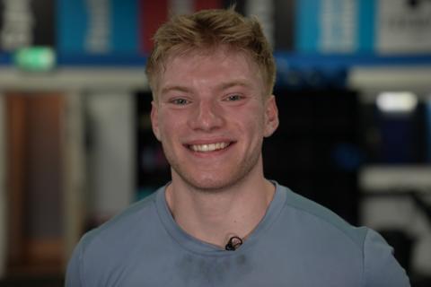 Close-up of a person in a light blue sports top, facing the camera and smiling. The background is softly blurred, showing an indoor gym. 