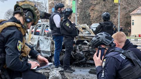 Six Ukrainian experts wearing helmets, and some of them wearing dark blue uniforms, work at the site of a drone attack in the Ukrainian city of Kharkiv. Behind them stands a wrecked car.