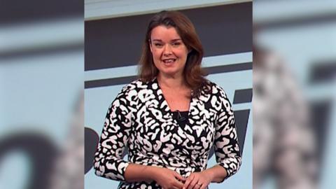 A woman with brown hair is wearing a black and white printed dress is standing in front of a screen that is projecting the weather forecast.