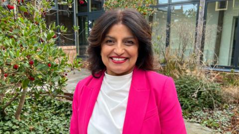 Charnjit Saranna smiles at the camera. She is in front of a green tree with red berries and is wearing a pink jacket and white shirt.