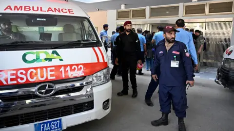 An ambulance is parked outside the hospital next to security staff and onlookers