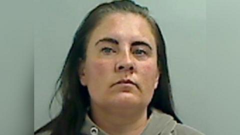 A woman with long dark hair and dark eyebrows looks at the camera in a police mugshot. She is wearing a grey hoodie.