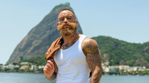 The image shows a single person standing outdoors in a bright, sunny setting with a well-known coastal landscape in the background.