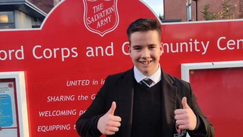 A boy in a black school uniform and tie smiles at the camera with his thumbs up. He is in front of a red Salvation Army sign.
