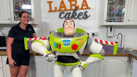 A woman standing next to large Buzz Lightyear cake. The woman has brown hair tied back and the cake is white and green with three small buttons which are blue, green and red and a large red button. The cake also has wings which are striped red and white.
