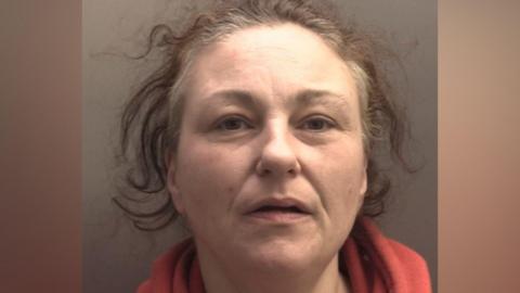 A woman in her 40s, with unkempt brown hair and wearing a red fleece, looks into the camera with a slight smile
