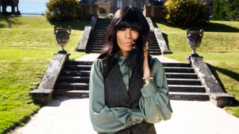 Claudia Winkleman standing in front of the steps leading up to the Traitors castle