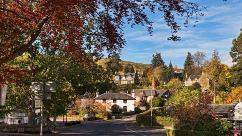 Strathpeffer village in the sunshine. There are a number of Victorian buildings and a large tree with red leaves.