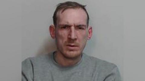 A police mugshot of a man with short brown hair and wearing a grey sweatshirt.