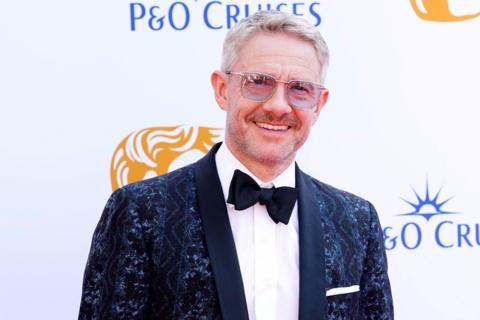 Actor Martin Freeman stands smiling at the camera. He is wearing a formal jacket with a blue paisley pattern worked into it,  a black bow tie and sunglasses