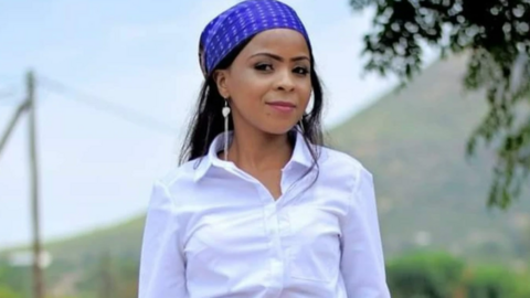 Keotshepile Naso Isaacs looking directly at the camera. She is wearing a white shirt. She is wearing a blue headband, which covers the front of long dark hair. She has long heart shaped earrings on and is standing in front of foliage with a blue sky in the background.