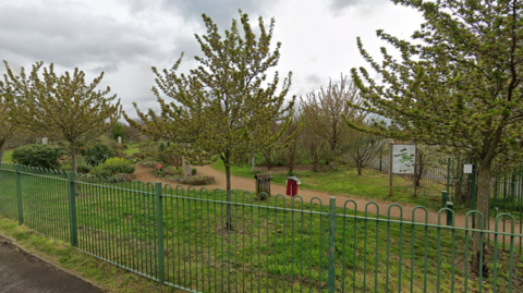 A street view image of a small grass park with small trees and a brown footpath running through it. The photo is taken from outside the park. There are metal railings on the edge of the park