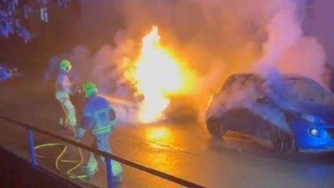 Blue car on fire, with firefighters with a hose trying to put the blaze out. 