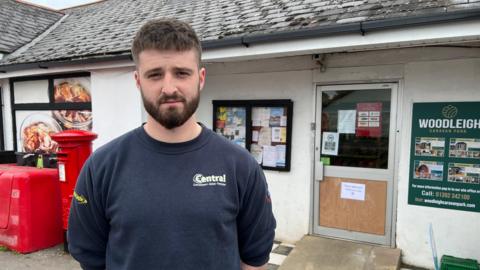 The shop manager stands outside his shop with a serious expression on his face. 