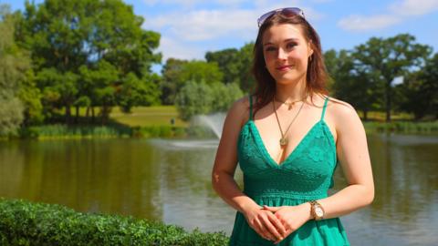 A woman with a green dress with her hands clasped is standing in front of a lake with a fountain and trees in the background