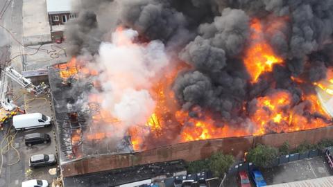 A large single storey building has a raging orange fire inside and thick black smoke rising from it.