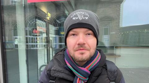 Lee Tyler - a man with a grey beanie, striped scarb and black coat looks directly at the camera
