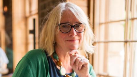 A woman with blonde hair and glasses looks at the camera and touches her chin. She is wearing a navy top, green cardigan, and a necklace of large green and brown circles, and is sat indoors near a window.