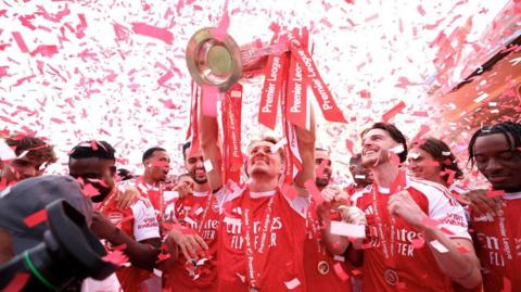  An Arsenal player lifts the Premier League trophy above his head as red and white confetti falls around him, with teammates in red shirts celebrating alongside.