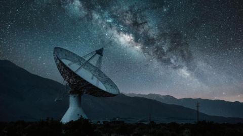 A large radio telescope dish stands in a dark landscape with low hills, set beneath a clear night sky filled with stars and the Milky Way band.
