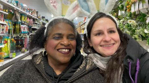 Two women wearing bunny ears made from wire and fluffy fabric. They are smiling and standing in a shop.