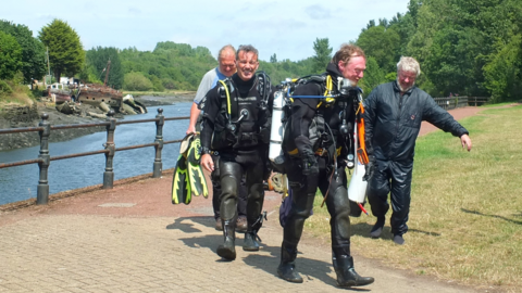 Two divers in full gear walking along the river bank after getting out of the River Wear. They are smiling. Two men are walking beside them and carrying equipment. 
Gary Bankhead is one of the divers. He is smiling and has short grey hair.