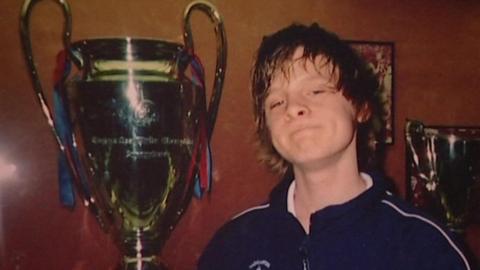 A large trophy with two handles, resembling the UEFA Champions League cup, is prominently displayed in the centre. The trophy has ribbons in blue and red attached to its handles. Joe stands to the right, he is wearing a dark sports jacket with a white zipper and a crest on the chest.