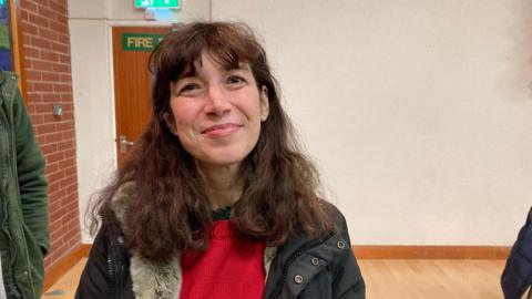 Manuela has wavy red-brown hair with a fringe. She is standing in a hall and is wearing a red jumper and black coat