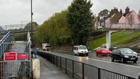 A partially demolished footbridge over a busy road and a sign saying footpath closed in the foreground 