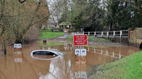 A silver car is almost completely submerged under flood water. A red sign next to the vehicle reads: "Do not cross when in flood." 