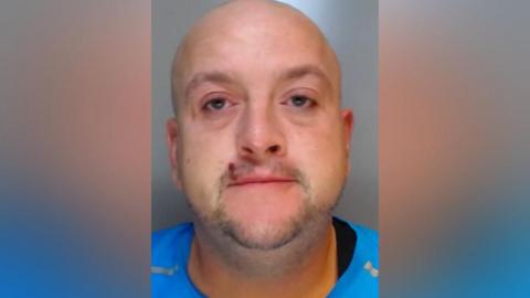 Mugshot of Steele. He is bald with faint stubble and a cut above his lip