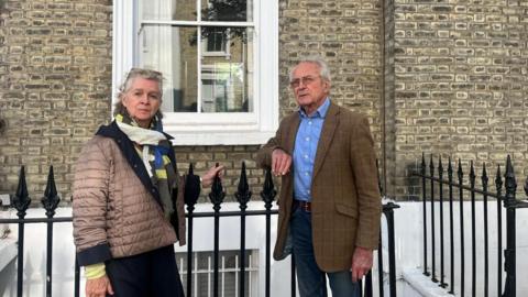 Two neighbouring residents standing beside a black iron railing outside a brick townhouse, with a window visible behind them.