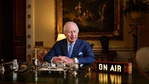 King Charles, wearing a suit, sitting behind a desk in Buckingham Palace with a painting behind