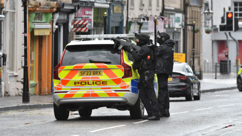Two police officers dressed in black are pointing guns at an upper window on a street from behind a marked police car