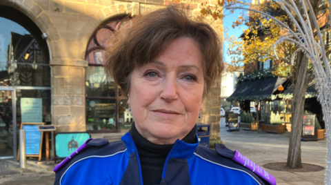 A woman with dark hair and wearing a blue top with two different shades of blue is looking at the camera.