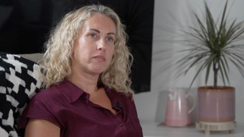 A woman with blonde curly hair and maroon dress sits in a sofa with a spiky pot plant behind her