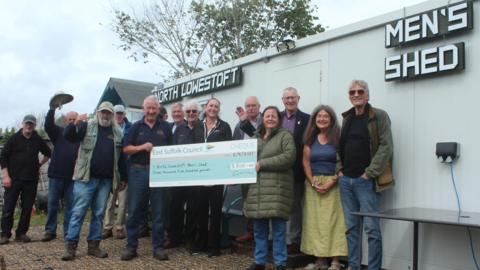 A group of men and women stand in front of a large shed smiling at the camera. Some of the people at the front hold a large cheque that has been donated to the group. One group member raises their hat in the air.