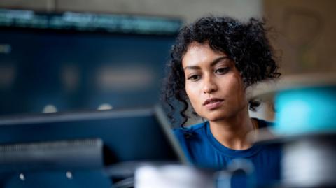A woman with dark hair works on laptop. She is wearing a blue top.