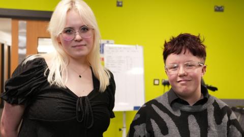 One student with long blonde hair and glasses and a shorter student with short dark hair and glasses standing next to each other in front of a whiteboard