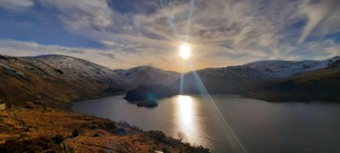 Sun shines in a partly cloudy sky above snow-capped mountains next to a lake