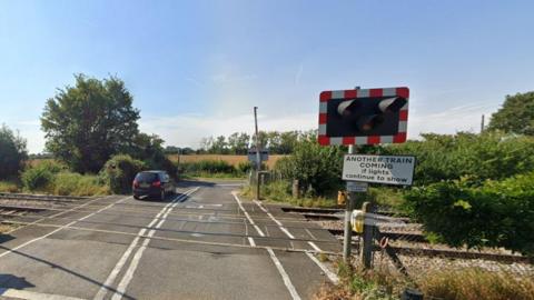 Sandy Lane level crossing on a sunny day. A car is seen travelling away from the crossing. A sign reads "Another train coming if lights continue to show".