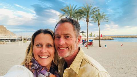 A couple on a beach. It is sunny and the sand is white. Behind them are some trees.
